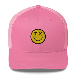 May include: Pink and black trucker hat with a yellow smiley face embroidered on the front. The smiley face has the letters "TX" for eyes.