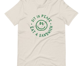 May include: A beige t-shirt with a green circle graphic that says "Sit in peace, eat a sandwich" with a smiley face in the center.