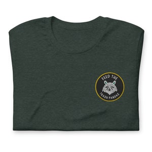 以下が含まれることがあります： 黒い円形の刺繍パッチがついたダークグリーンのTシャツ。パッチには「ゴミパンダに餌をやる」と書いてあり、その中にアライグマがいます。