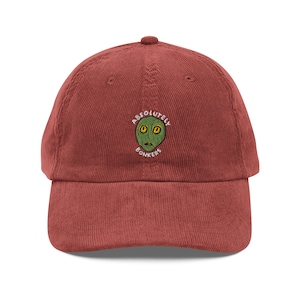Puede incluir: Gorra de béisbol de pana color ladrillo. La gorra presenta un rostro alienígena bordado con el texto "ABSOLUTELY BONKERS" en un diseño circular. La gorra tiene una visera curva y una correa ajustable.