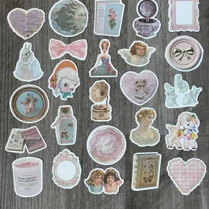 May include: A selection of vintage-style stickers with diverse designs, such as cherubs, a lamb, a doll, a bow, a moon face, a heart, a book, and a candle. The stickers have a soft, pastel colour palette with pink, blue, and cream tones.