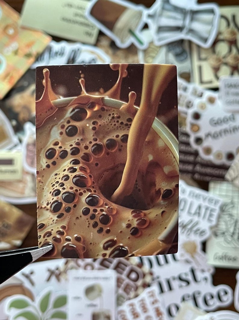 May include: A rectangular sticker featuring a close-up of a light brown coffee beverage being poured into a cup, creating a splash and numerous dark bubbles on the surface. The liquid is a creamy tan color with deep brown droplets and foam.