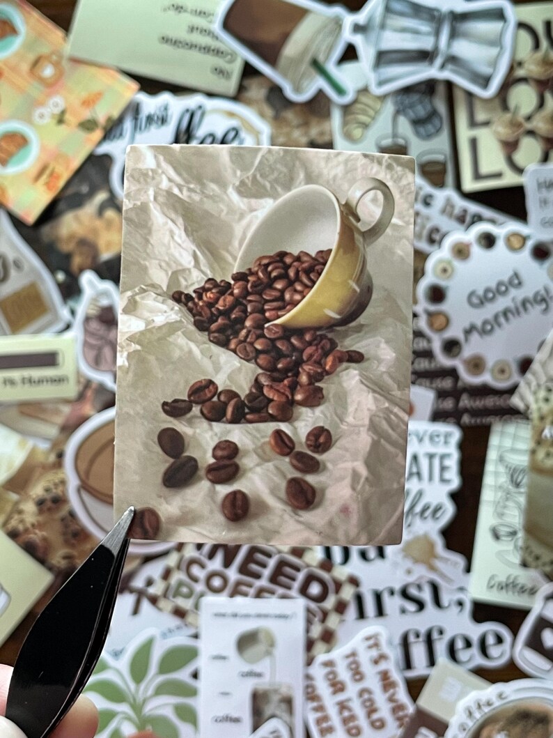 May include: A rectangular sticker featuring a cream-colored ceramic coffee cup tipped on its side, spilling dark roasted coffee beans onto a textured, crumpled off-white paper background. The image captures the beans scattered across the surface.