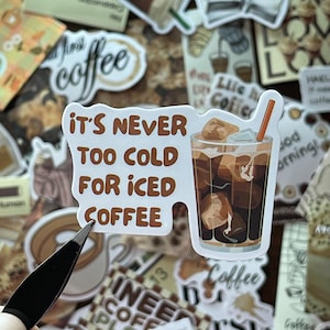 May include: A die-cut vinyl sticker featuring a glass of iced coffee with ice cubes and an orange straw, accompanied by the brown text "IT'S NEVER TOO COLD FOR ICED COFFEE" arranged in a block format to the left of the beverage illustration.