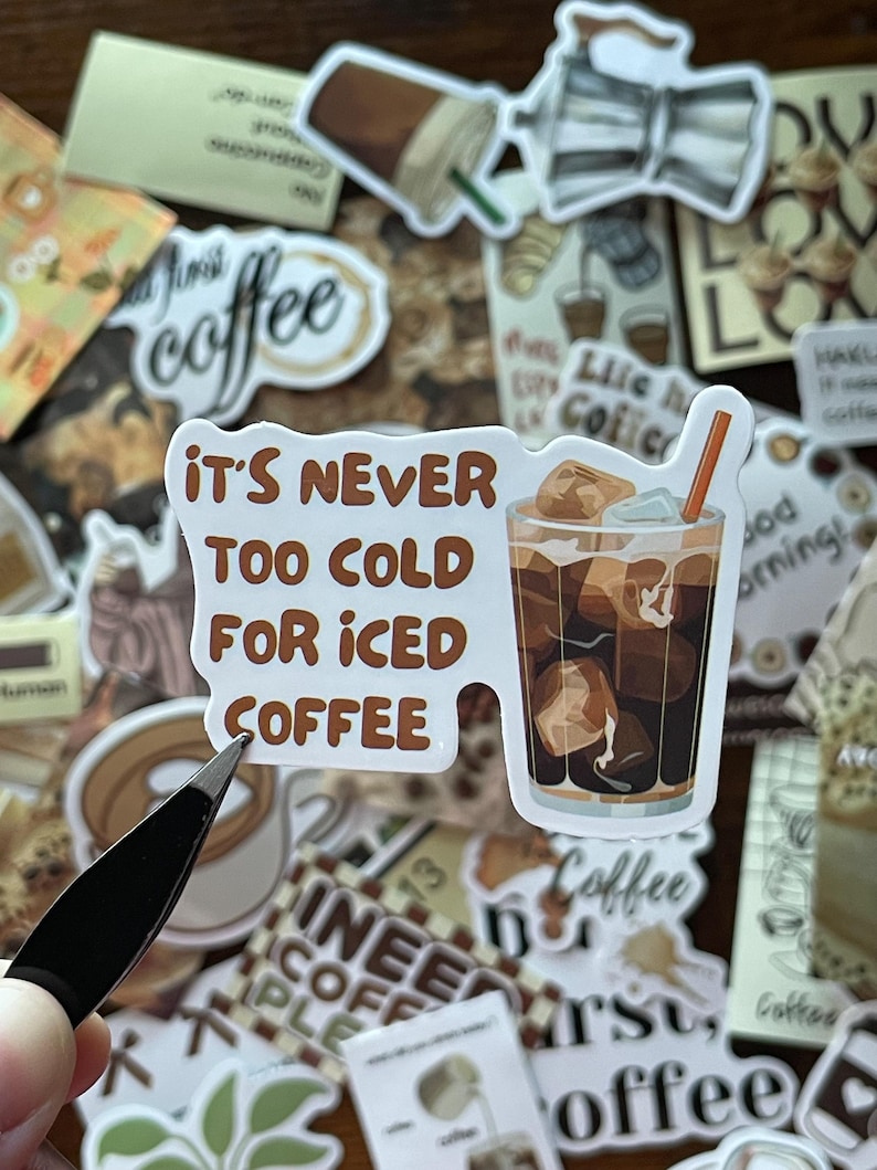 May include: A die-cut vinyl sticker featuring a glass of iced coffee with ice cubes and an orange straw, accompanied by the brown text "IT'S NEVER TOO COLD FOR ICED COFFEE" arranged in a block format to the left of the beverage illustration.
