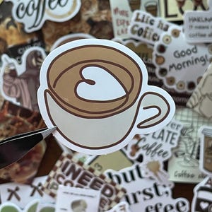 May include: A die-cut vinyl sticker featuring a stylized illustration of a light beige coffee cup filled with brown liquid. The surface of the coffee displays a white heart-shaped latte art design outlined in dark brown, set against a background.