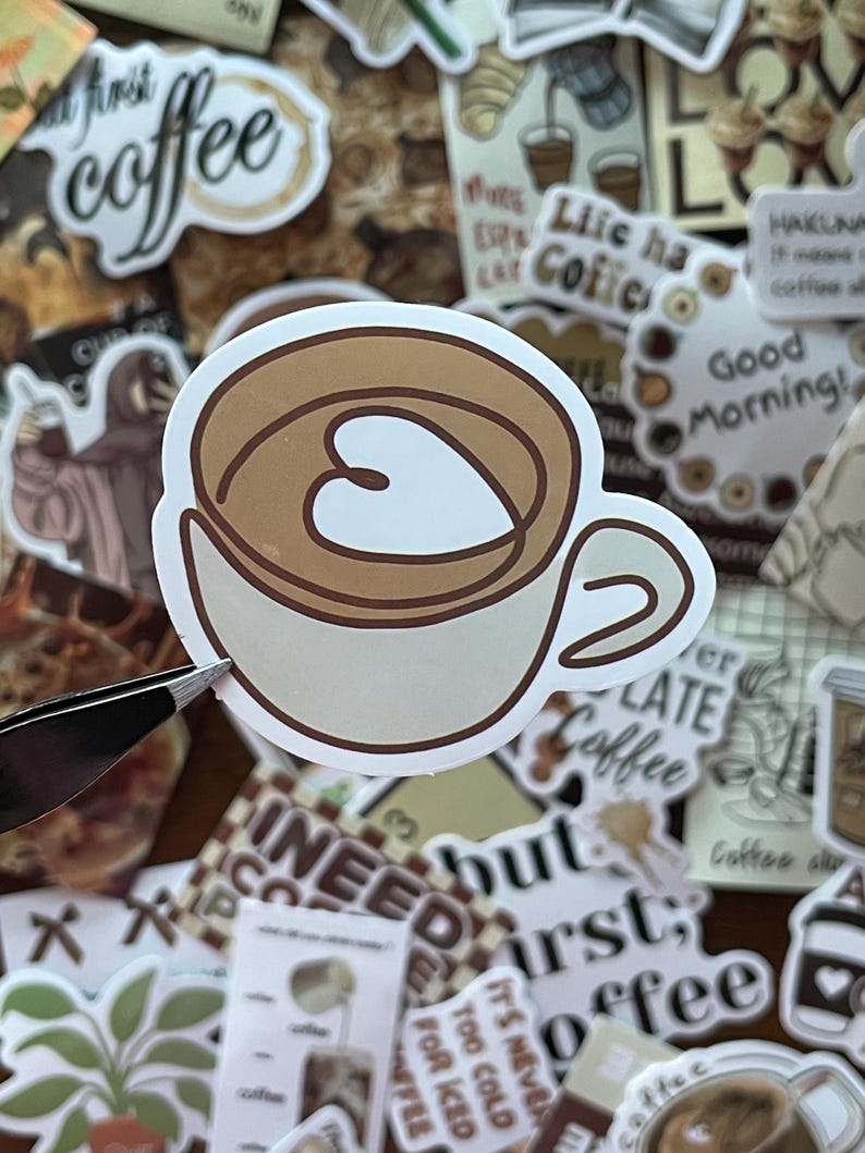 May include: A die-cut vinyl sticker featuring a stylized illustration of a light beige coffee cup filled with brown liquid. The surface of the coffee displays a white heart-shaped latte art design outlined in dark brown, set against a background.
