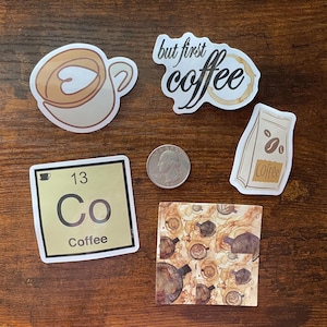 May include: A collection of five coffee-themed stickers including a latte cup, a script text sticker reading but first coffee, a coffee bean bag, a periodic table element square labeled 13 Co Coffee, and a square with coffee ring stains and a US quarter.