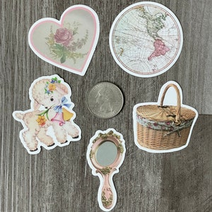 May include: A selection of vintage stickers, including a heart with a rose, a world map, a lamb, a hand mirror, and a picnic basket. The stickers have white borders and are displayed on a grey wooden surface.