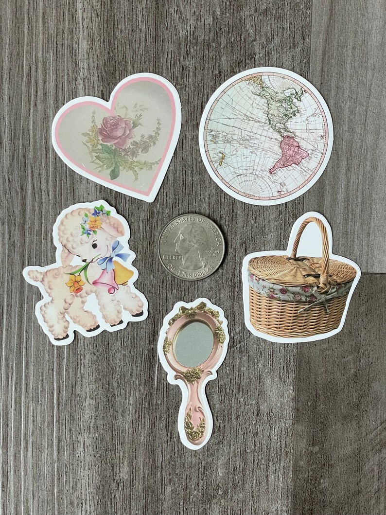 May include: A selection of vintage stickers, including a heart with a rose, a world map, a lamb, a hand mirror, and a picnic basket. The stickers have white borders and are displayed on a grey wooden surface.