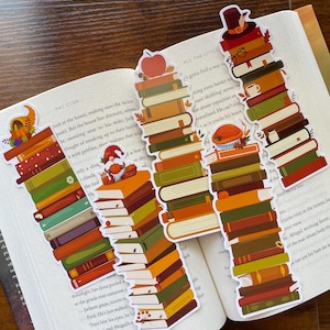May include: Four vertical bookmarks featuring illustrations of stacked books in autumn colors. The designs include a cornucopia, a gnome with a pumpkin, a red apple, a pilgrim hat with glasses, and a pie, all decorated with fall leaves and foliage.