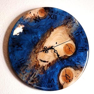 May include: Round wall clock with a blue resin and wood design. The clock face features a natural wood grain center with blue resin accents and black Roman numerals. The clock hands are black.
