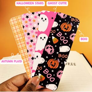 May include: Four rectangular bookmarks featuring distinct patterns: an orange and white autumn plaid, a pink background with black and white stars labeled HALLOWEEN STARS, a pink background with white ghosts labeled GHOST CUTTIE, and a black background with pumpkins, candy corn, and the text BOO.