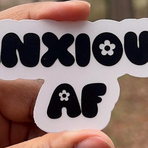May include: White sticker with black text that reads "ANXIOUS AF" with a small white flower in each letter.