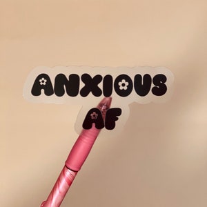 May include: A clear sticker with black text that reads "ANXIOUS AF" in a bubble font. The sticker has small white flower accents. A pink pen is holding the sticker.
