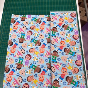 Op de afbeelding: Blauwe stof met een herhalend patroon van cartoonkinderen met verschillende huidtinten en haarkleuren. De kinderen dragen kleurrijke kleding en houden elkaar vast. De stof is perfect voor knutselprojecten.