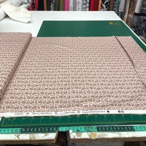 Könnte beinhalten: Stoffrolle mit sich wiederholendem Blumenmuster in Rosa und Braun auf cremefarbenem Hintergrund. Der Stoff, mit der Bezeichnung "New Canterbury", liegt auf einer grünen Schneidematte. Ideal für Näh- und Bastelprojekte.