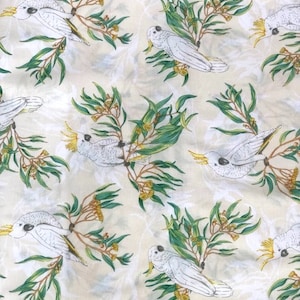 Op de afbeelding: Een witte stof met een herhalend patroon van witte kaketoes die op groene eucalyptus takken zitten met gele bloemen.