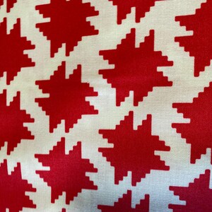 May include: A close-up of a fabric featuring a repeating pattern of red starburst shapes against a white backdrop. The starbursts have a pixelated design, creating a bold graphic. The fabric appears to be cotton or a similar material.