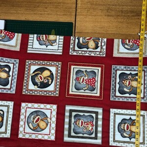 May include: Red fabric with a repeating pattern of monkeys wearing different outfits. The monkeys are in white frames with a polka dot background. The fabric has a white border with the text "Dane Design 2020 for QT fabrics www.QTfabrics.com".