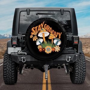 May include: Black spare tire cover with a colorful design featuring mushrooms, flowers, and the words "Stay Groovy" in orange and yellow. The cover is mounted on the back of a black Jeep, set against a backdrop of a road and mountains.