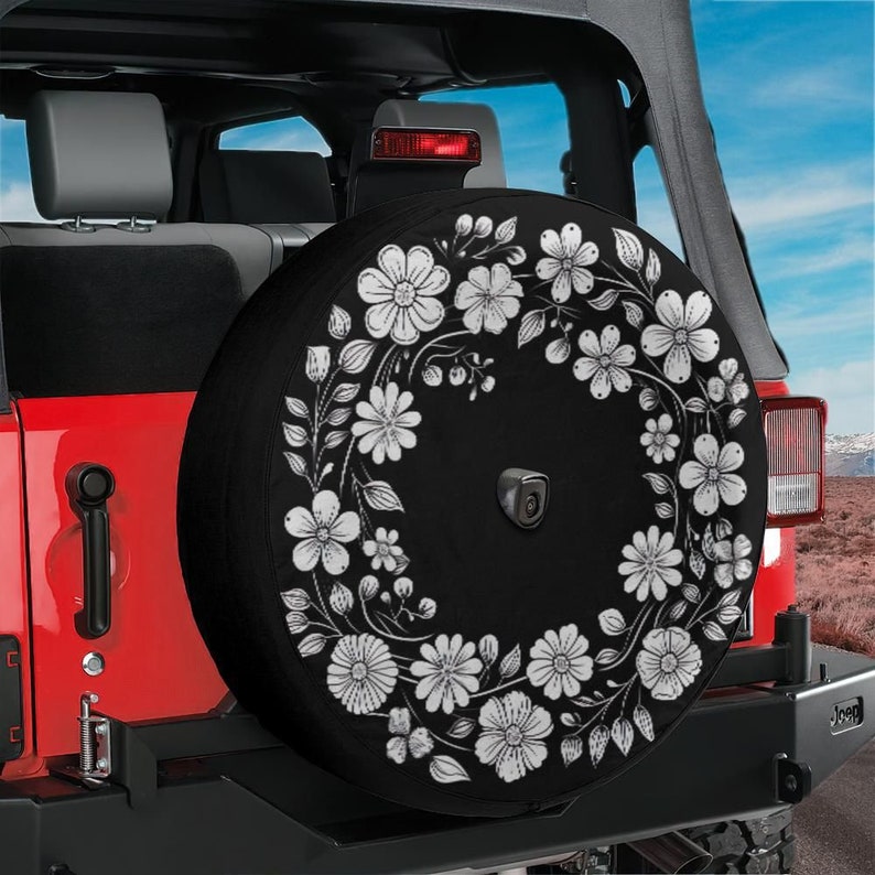 May include: Black spare tire cover with a white floral design. The cover features a wreath of white flowers and leaves on a black background.
