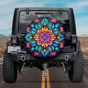 May include: A black Jeep with a colorful spare tire cover featuring a vibrant mandala design. The mandala has a symmetrical pattern with shades of blue, orange, purple, and green. The vehicle is parked on a road with a mountain backdrop.