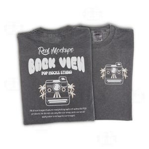 May include: Two charcoal grey t-shirts. One displays "Real Mockups BACK VIEW POP MOCKS STUDIO" with a camera graphic and palm trees. The other shirt has a camera graphic with palm trees. The shirts are laid flat.