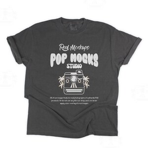 May include: Dark grey t-shirt with rolled sleeves. The shirt displays the text "Real Mockups POP MOCKS STUDIO" in white, and a camera graphic. Below the graphic is text that reads "All of our images feature real photographs of authentic POD products..."
