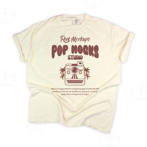 May include: A cream-coloured t-shirt with rolled sleeves. The shirt displays a brown graphic of a vintage camera with palm trees and the text "Real Mockups POP MOCKS STUDIO". Additional text is below the graphic.