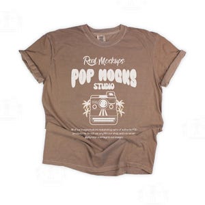 May include: A brown t-shirt with rolled sleeves. The shirt displays the white text "Real Mockups POP MOCKS STUDIO" and a camera graphic. The t-shirt is made of a soft material.