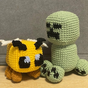 Pode incluir: Um brinquedo de peluche de abelha de crochê amarelo e marrom com olhos pretos e um brinquedo de peluche de creeper de crochê verde com olhos pretos e uma boca preta.