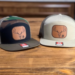 May include: Two trucker hats with a brown leather patch featuring a deer skull with antlers. One hat is camouflage with a black crown and a green brim. The other hat is beige with a gray mesh back.