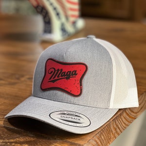 May include: A gray and white trucker hat with a red and black patch that says "Maga" on it. The hat has a white mesh back and a gray front. The hat has a label that says "The Authentic Snapback" on it.