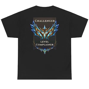 May include: Black t-shirt featuring a graphic design with the words "CHALLENGER" and "LEVEL COMPLAINER" above and below a stylized emblem. The emblem has blue and gold accents.