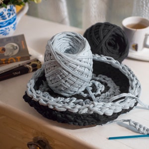 May include: A close-up of two balls of yarn, one light blue and one dark gray, with a crocheted basket and a crochet hook. The yarn is made of t-shirt yarn.