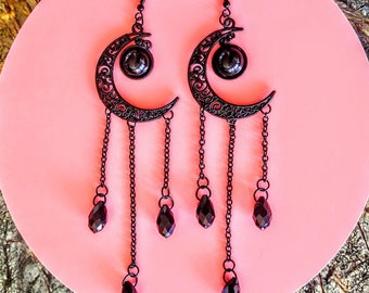 May include: A pair of black metal earrings featuring a crescent moon design with dangling chains and teardrop-shaped dark red crystals. The earrings have a hook closure and are displayed on a pink surface.