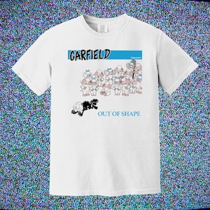 May include: White t-shirt featuring the word "GARFIELD" in blue and a cartoon image of Garfield and other characters. The text "OUT OF SHAPE" is below the cartoon image. The shirt is a crew neck.