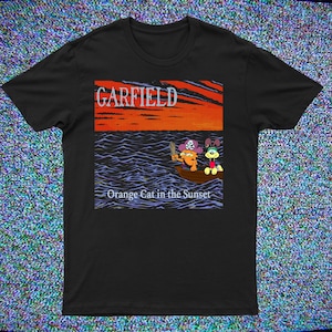 May include: Black t-shirt featuring a Garfield graphic. The design shows Garfield as a pirate in a boat on the ocean at sunset, with the text "GARFIELD" above and "Orange Cat in the Sunset" below. The shirt is a classic crew neck.