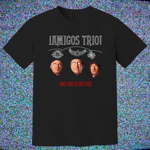 May include: Black t-shirt with the text "¡AMIGOS TRIO!" above three illustrated faces. Below the faces, the text reads "FROM HERE TO SANTA POCO". The design includes white line art of suns, an eagle, and snowflakes.