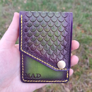 May include: A handcrafted leather wallet with a unique scale pattern in shades of green and purple. The wallet features a green pocket with the word "MAD" embossed in the leather, and is secured with a gold-colored button. Yellow stitching adds detail.
