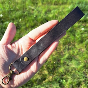 Puede incluir: Una correa de muñeca de cuero negro con un cierre giratorio y un remache de color latón. La correa se sostiene sobre un fondo de hierba verde.