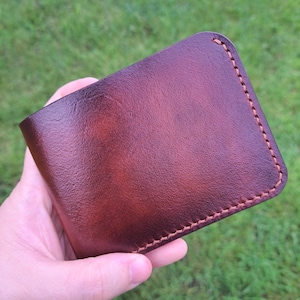 May include: A brown leather wallet with a reddish-brown hue. The wallet has a stitched border in a lighter brown thread. The leather appears smooth and has a slight sheen. The wallet is held in a hand against a blurred green background.