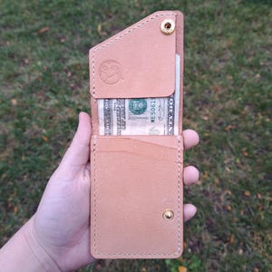 May include: A light brown leather wallet with a snap closure and a gold-coloured rivet. The wallet is open, revealing several US dollar bills. The wallet has a small embossed design on the front.