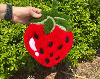 May include: A handmade strawberry-shaped rug with a vibrant red body, green leaves, and black seed details. The rug has a fluffy, textured appearance, and a white highlight. The rug is held in front of a green bush.