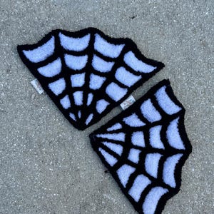May include: Two black and white spiderweb-shaped rugs. The rugs have a black border and a white web design. The rugs are placed on a gray concrete surface. The rugs are likely used as decorative items or floor mats.