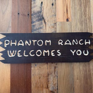 May include: A dark brown wooden sign with the words "PHANTOM RANCH WELCOMES YOU" carved in white. The sign has a jagged edge design and is mounted on a wooden plank background.