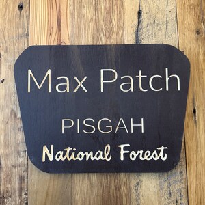 May include: A dark brown wooden sign with rounded edges and white text that reads "Max Patch Pisgah National Forest".
