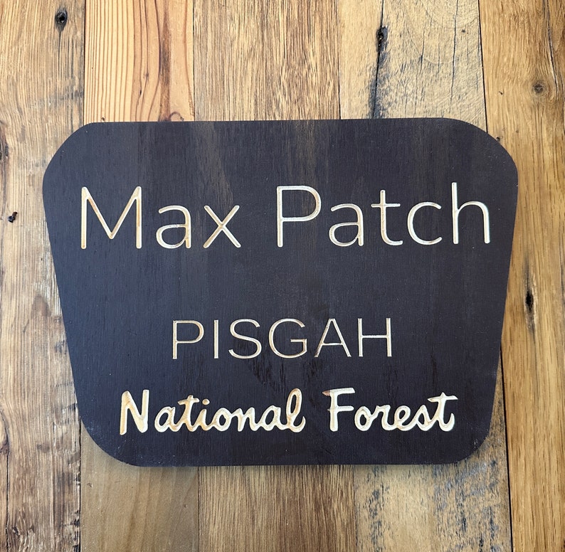 May include: A dark brown wooden sign with rounded edges and white text that reads "Max Patch Pisgah National Forest".
