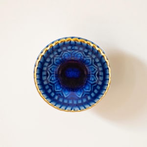 May include: A small, round, blue ceramic dish with a gold rim and a detailed mandala pattern. The dish is perfect for holding small items like jewelry or trinkets.